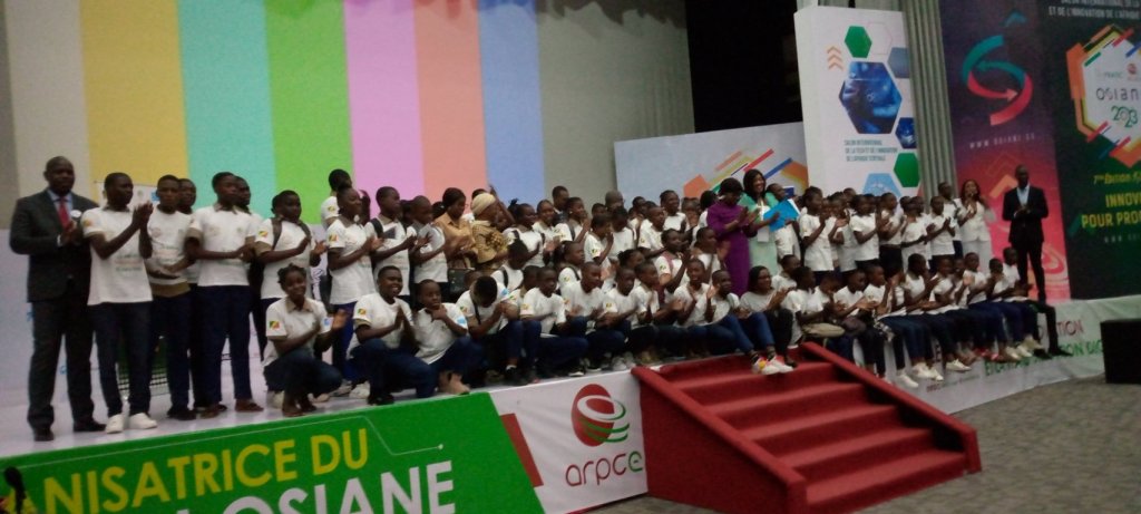 Un groupe d'élèves en t-shirts blancs applaudissant sur scène lors de l'atelier du Salon OSIANE 2025 à Brazzaville, entourés d'intervenants et d'un décor coloré.