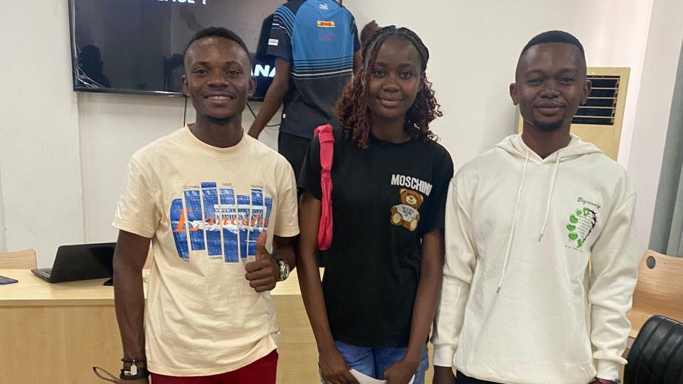 Trois étudiants posent ensemble dans une salle, avec un tableau en arrière-plan affichant le mot 'BIENVENUE'. L'un d'eux porte un t-shirt beige, un autre un t-shirt noir avec un motif amusant, et le troisième un sweat à capuche blanc.