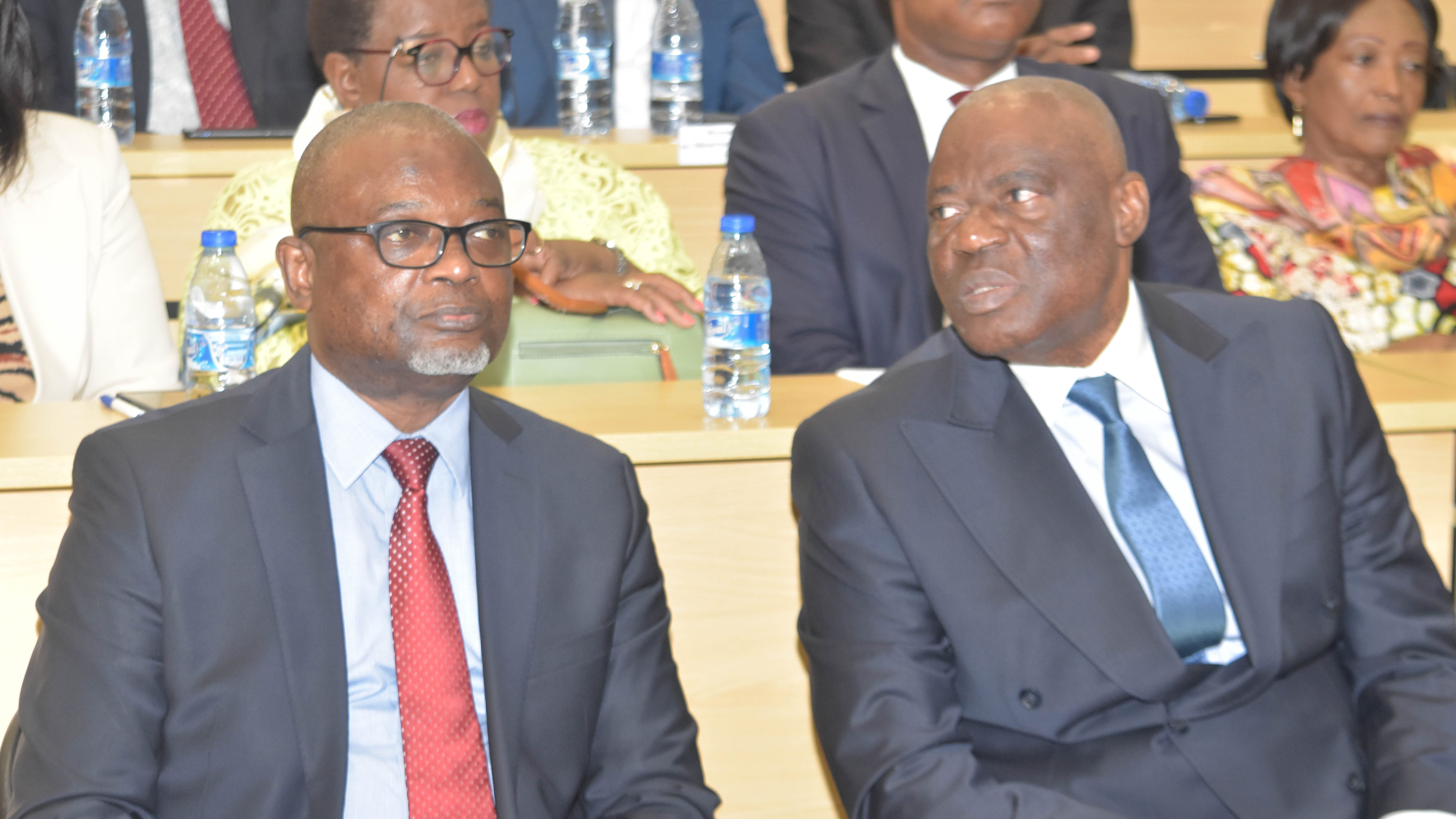 Deux hommes en costume assis sur des bancs dans une salle, avec d'autres participants visible en arrière-plan.