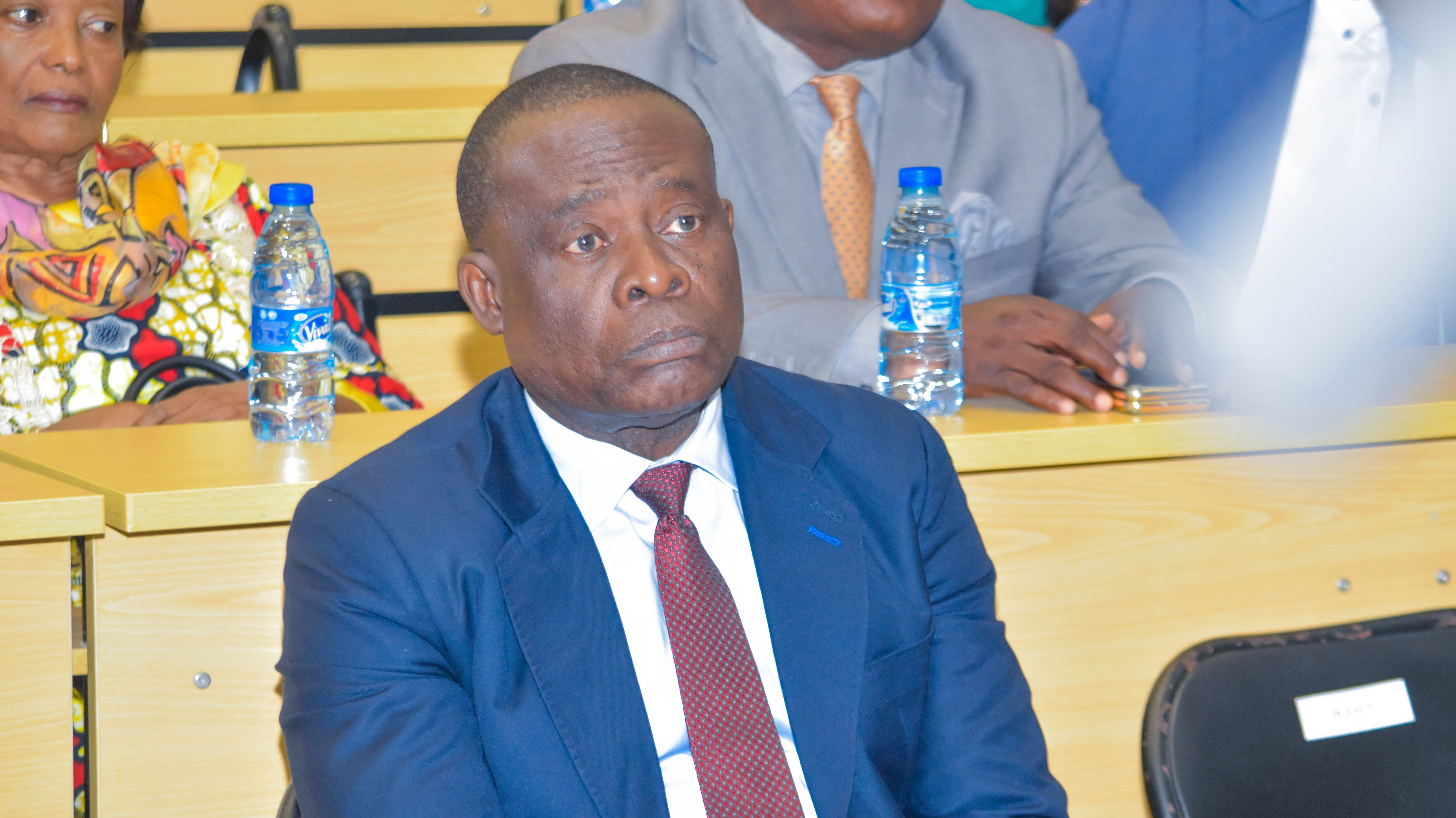 Un homme en costume assis, regardant attentivement lors d'un événement. Derrière lui, d'autres participants sont également présents, et des bouteilles d'eau sont visibles sur les tables.