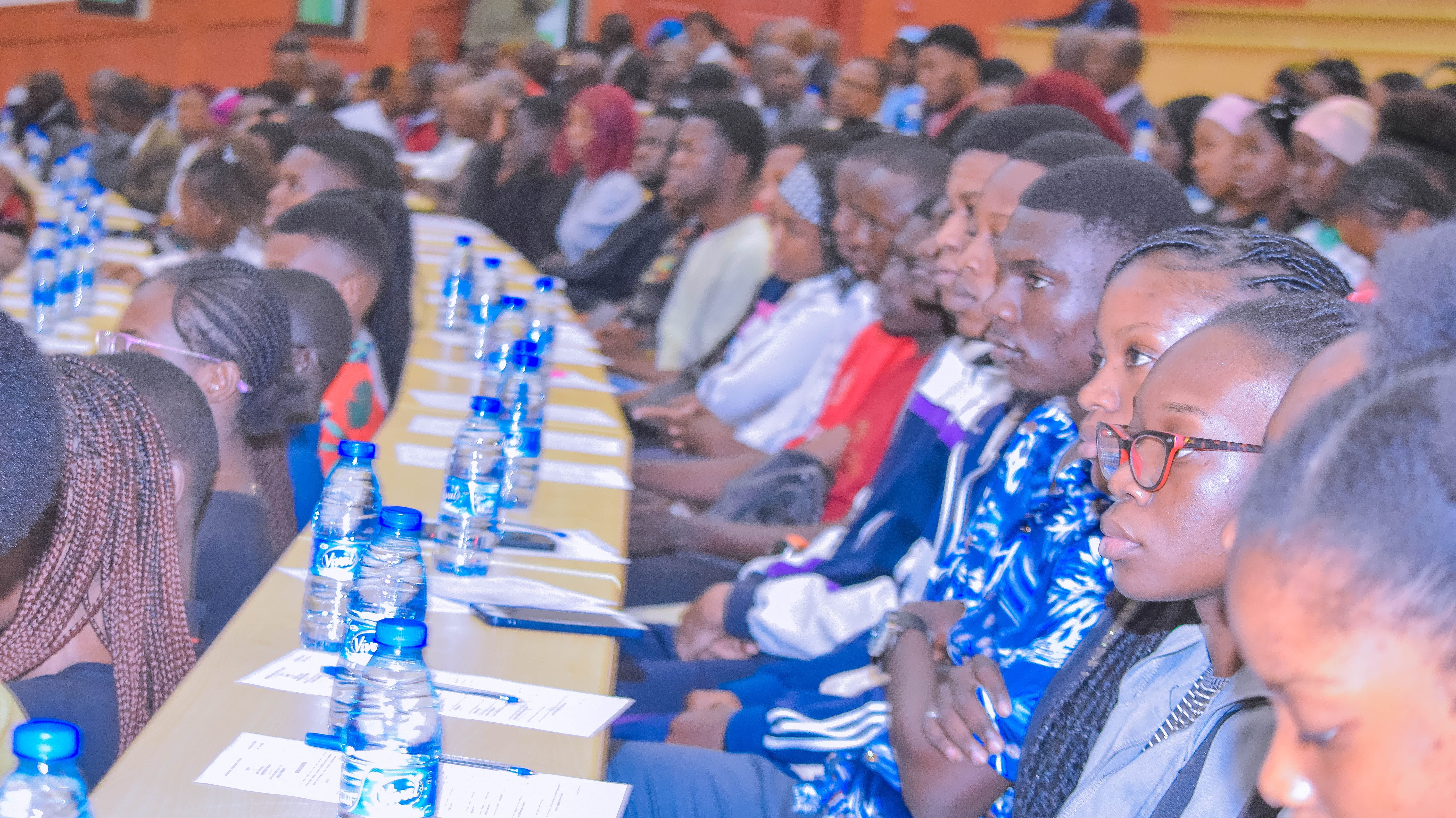 Un groupe d'étudiants attentifs lors du Salon de l'Information et de l'Orientation Universitaire à Brazzaville, avec des bouteilles d'eau et des documents sur les tables.