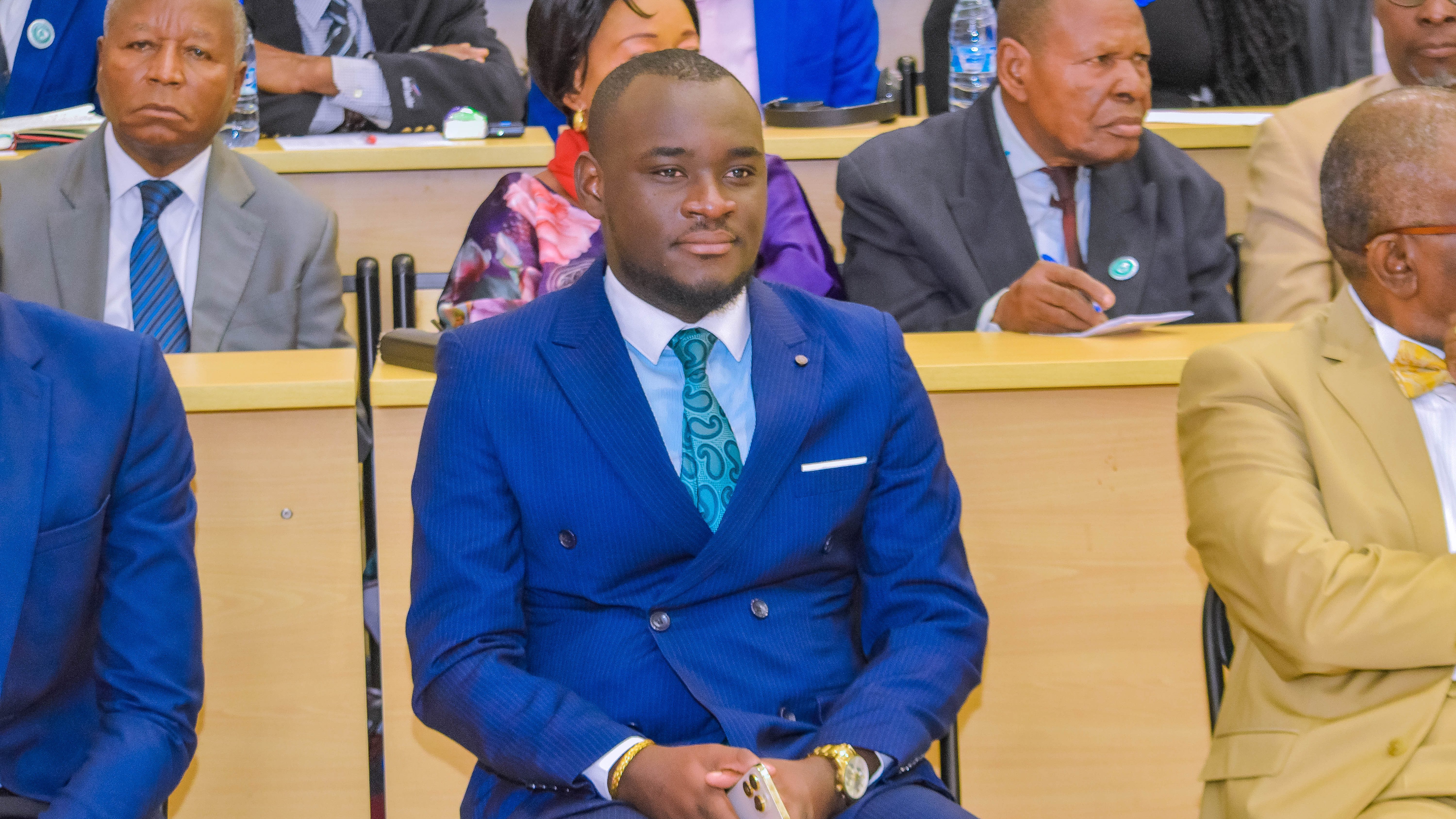 Un jeune homme assis dans un auditorium, portant un costume bleu, écoute attentivement, entouré par d'autres personnes au premier plan.
