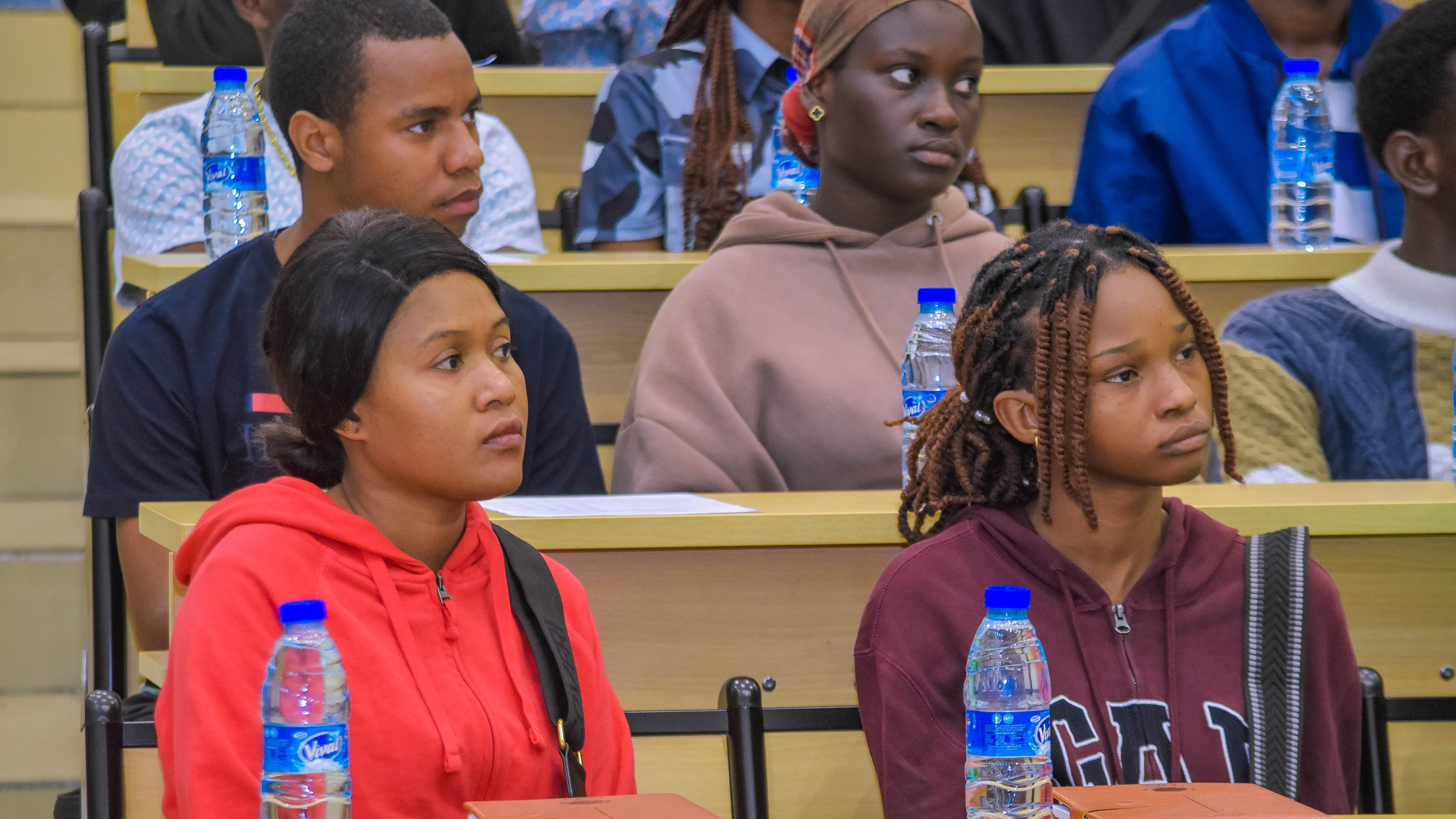 Des étudiants attentifs assistent à une séance d'information sur l'orientation universitaire, avec des bouteilles d'eau et des cahiers devant eux.