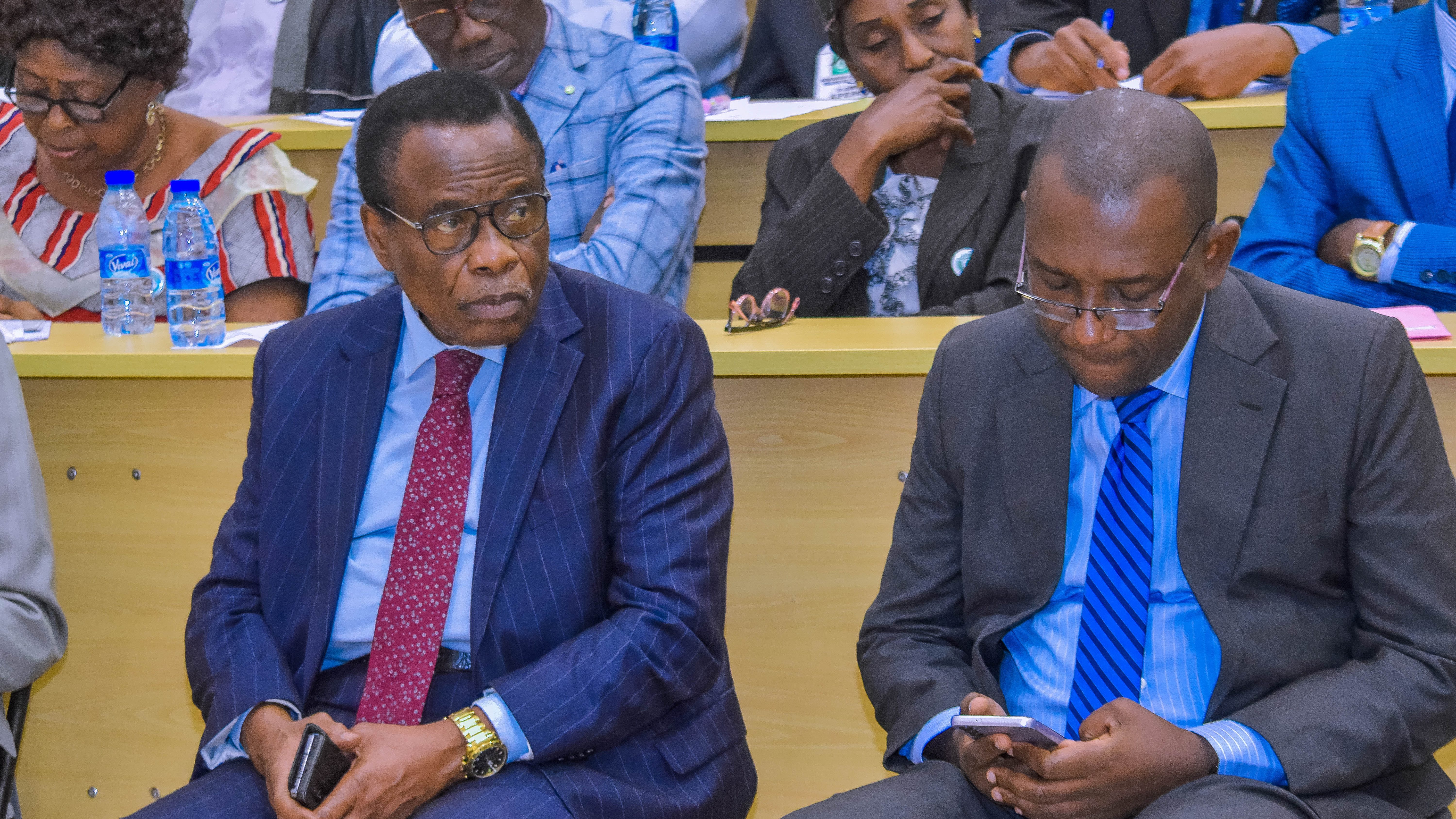 Deux hommes assis dans une salle de conférence, l'un portant une veste à rayures et une cravate rouge, l'autre en costume sombre avec une chemise à rayures bleues, concentrés sur leurs échanges.