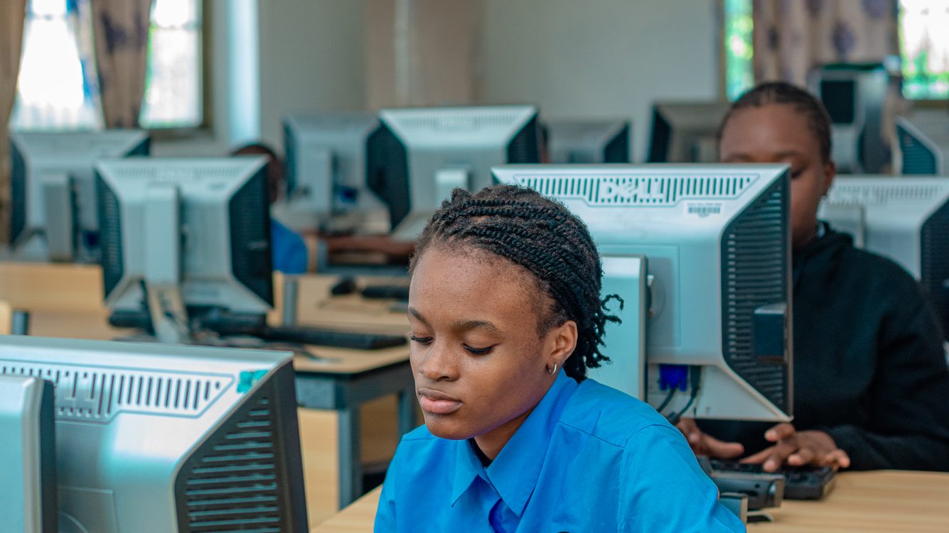 Un élève assis devant un ordinateur dans une salle de classe d'informatique, portant une chemise bleue, concentré sur son travail.