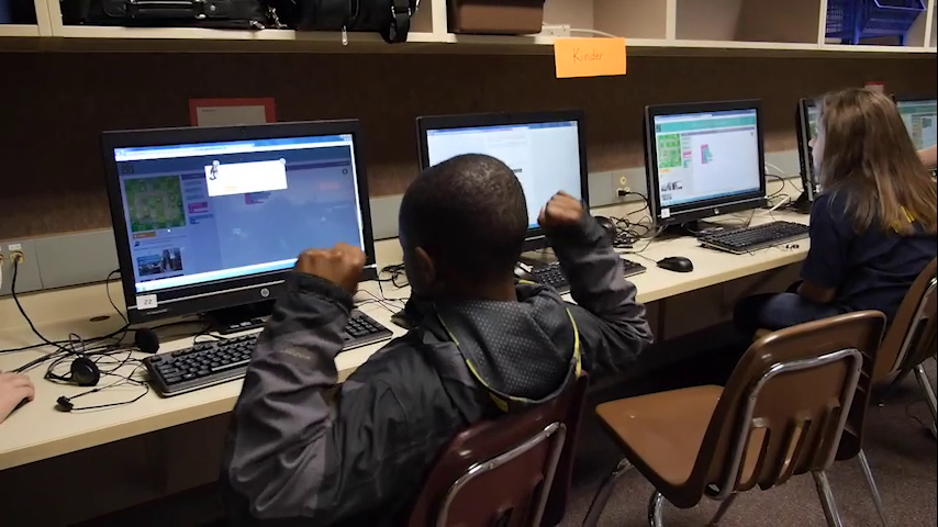 Un élève levant les bras en signe de victoire devant un ordinateur pendant un cours de programmation, avec d'autres étudiants à proximité utilisant des ordinateurs.