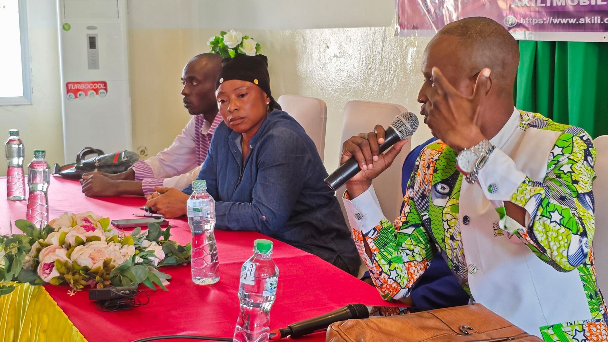 Une discussion lors d'un événement avec trois intervenants assis à une table, entourés de bouteilles d'eau et de fleurs. L'un des intervenants parle dans un microphone, tandis que les autres écoutent attentivement.