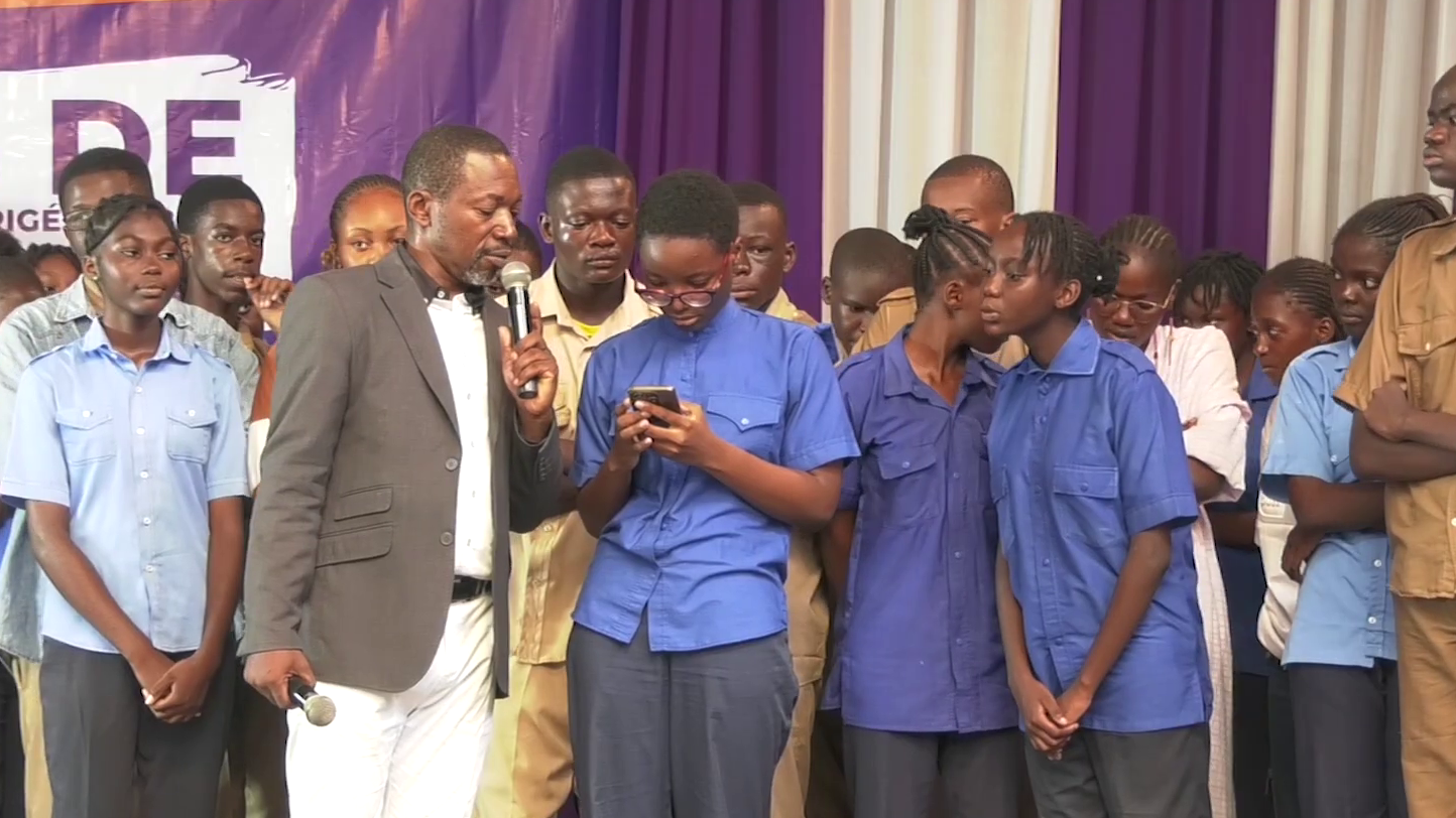 Cérémonie de remise des certificats pour les meilleurs élèves avec un présentateur parlant dans un micro, entouré de jeunes étudiants en uniforme.